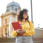 Emily J., United States Smiling female student with curly hair and glasses holding books on campus steps.