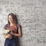 Aisha R., Canada Young woman holding books standing in front of a chalkboard filled with mathematical formulas.